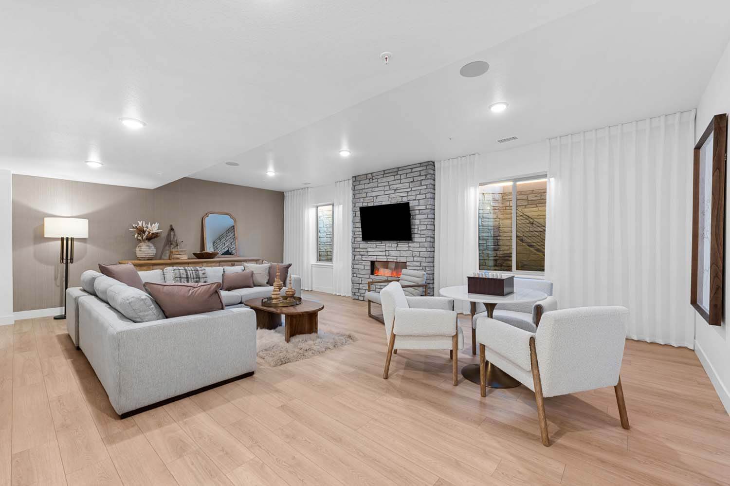 The rec room in the developed basement has a tan wallpapered accent wall, hardwood floors, a gas fireplace with a floor-to-ceiling stone surround and an oversized window on either side.