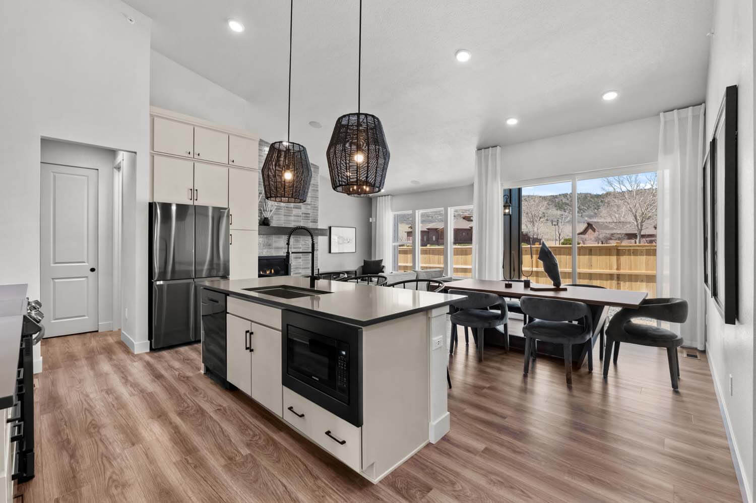 The open-concept floorplan has the kitchen, with its central island, looking to the dining area, which is adjacent to the great room with its vaulted ceiling. The space has white walls and hardwood floors.