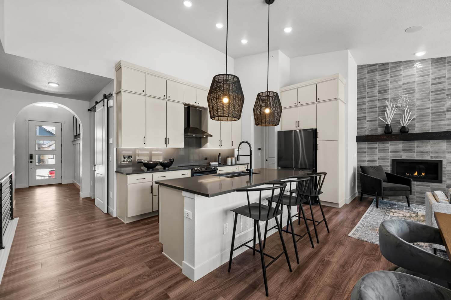 In the kitchen, there are two pendant lights above the island, dark quartz counters, beige cabinets, stainless steel appliances, and opens to a hallway leading to the foyer. The kitchen looks to the great room and dining room, and the space has a vaulted ceiling and hardwood floors.