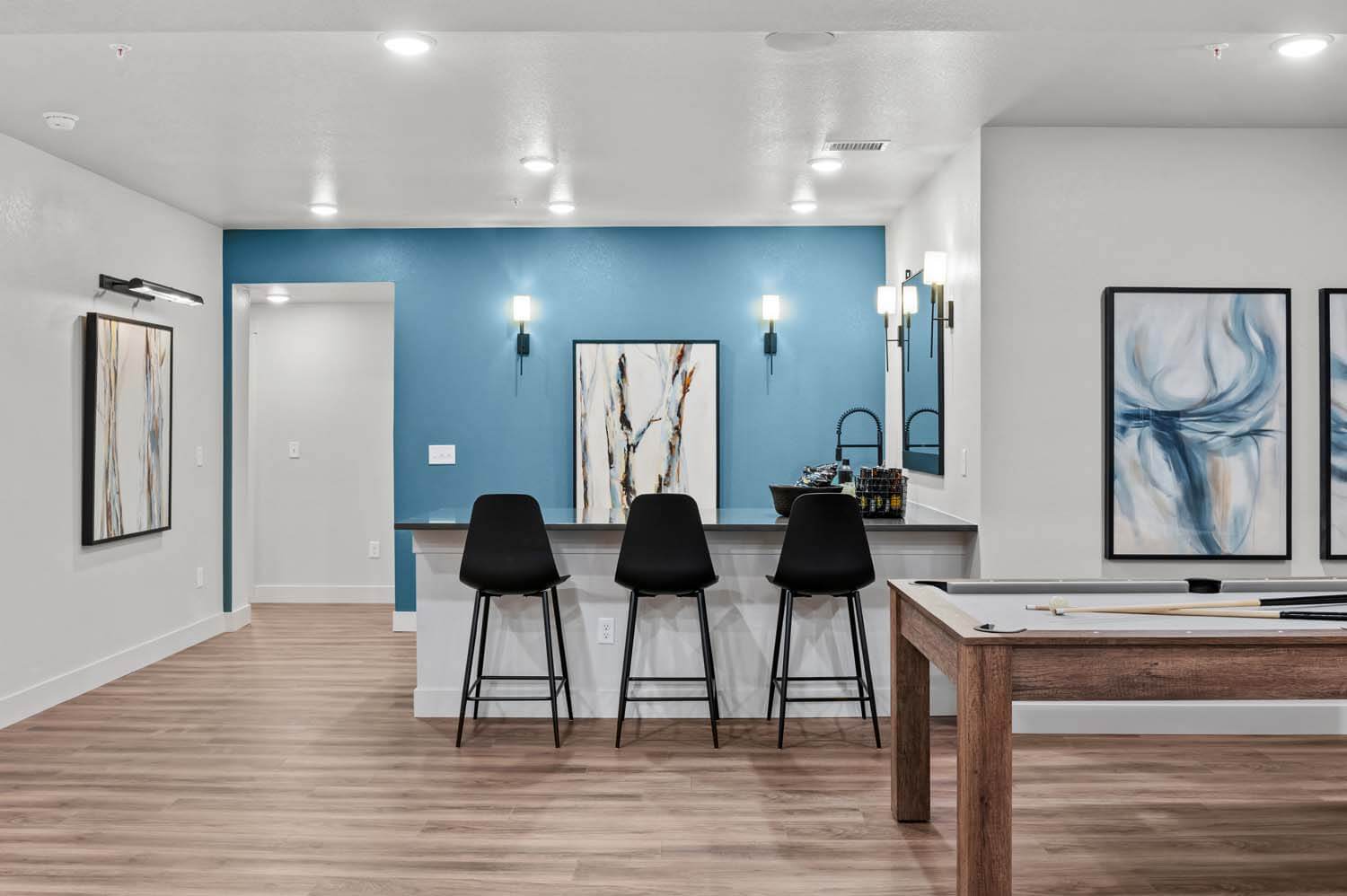 The wet bar area in the developed basement has a blue accent wall, hardwood floors, dark quartz countertops with a sink, beige cabinets and an island that easily fits three stools. There is an open space next to it that is ideal for a pool table.
