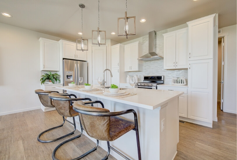A kitchen with hardwood floors, an island with three trendy pendant lights hanging over it, white quartz countertops and cupboards, a glossy white subway tile backsplash and stainless steel appliances.