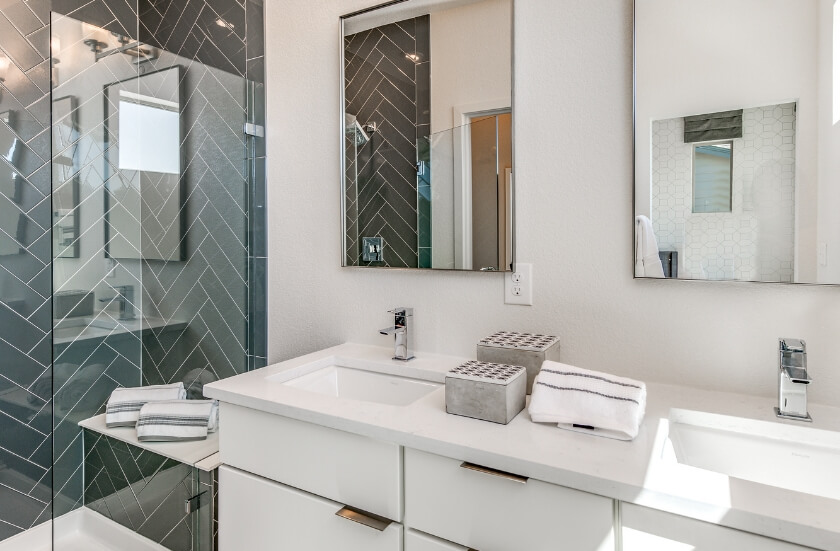 A bathroom with a double vanity with white counters and cabinets, mirrors above each rectangular undermount sink and grey herringbone-patterned tile in the spacious walk-in shower.